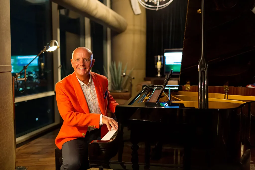 Lord Russel Baker at the piano with an orange jacket.